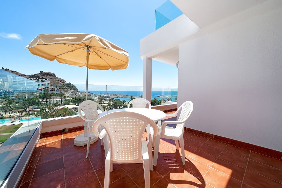 A balcony with white chairs and a table, a beige umbrella, and a view of the sea and buildings.