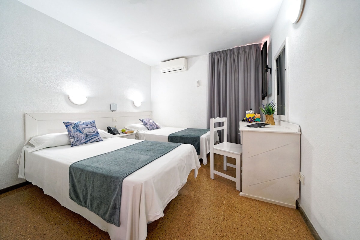 A hotel room with two beds, a desk, a chair, a mirror, and an air conditioning unit.