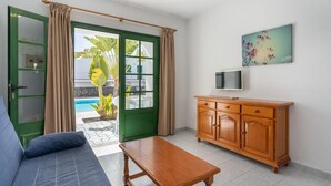 A room with a sofa, a wooden table, a TV on a wooden cabinet, and a door leading to a pool area. - Aloe (Puerto del Carmen)
