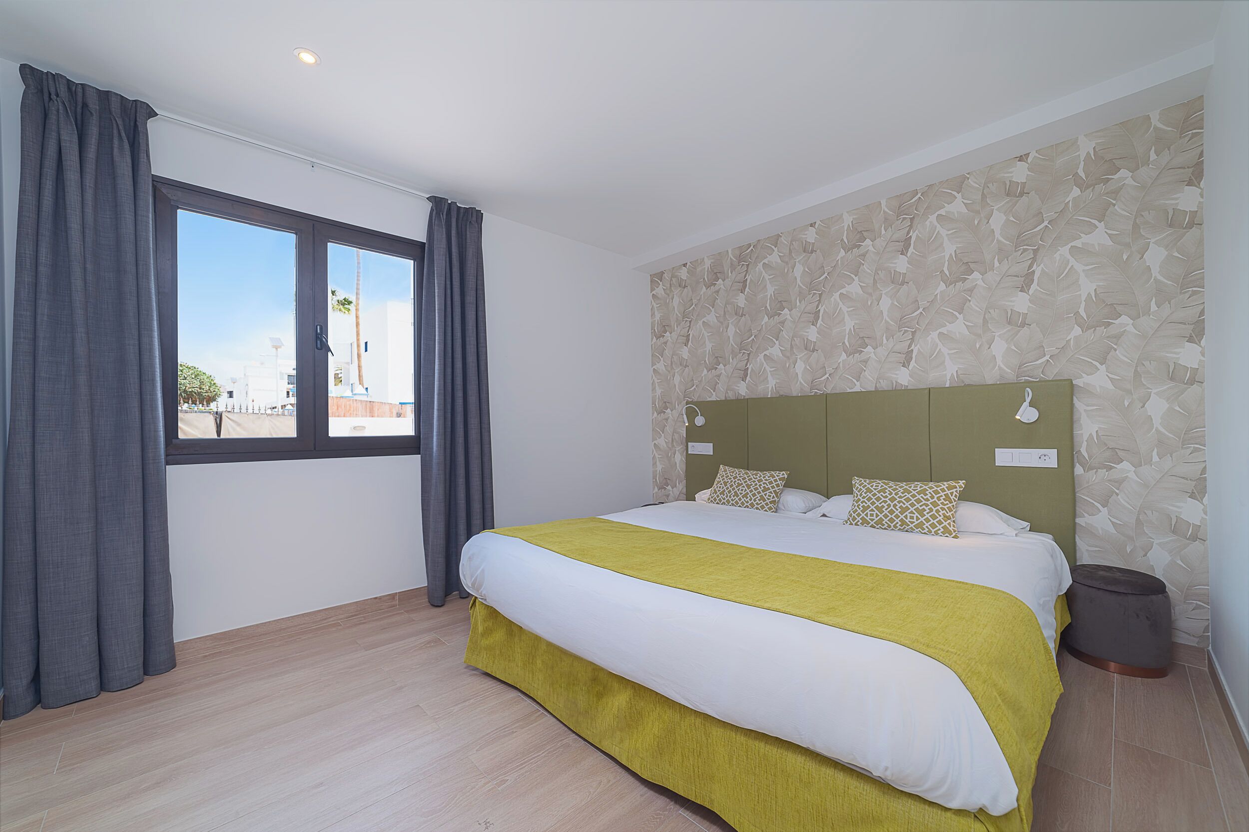 A hotel room with a large bed, a headboard, a window with curtains, and a patterned wall.