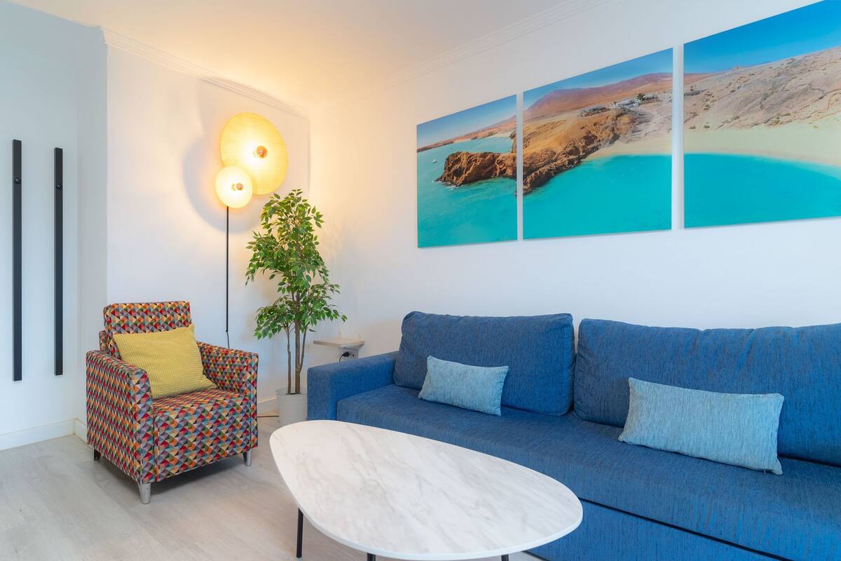 Un salon avec un canapé bleu, un fauteuil à motifs, une table basse blanche et un lampadaire. Trois tableaux représentant des paysages côtiers sont accrochés au mur.