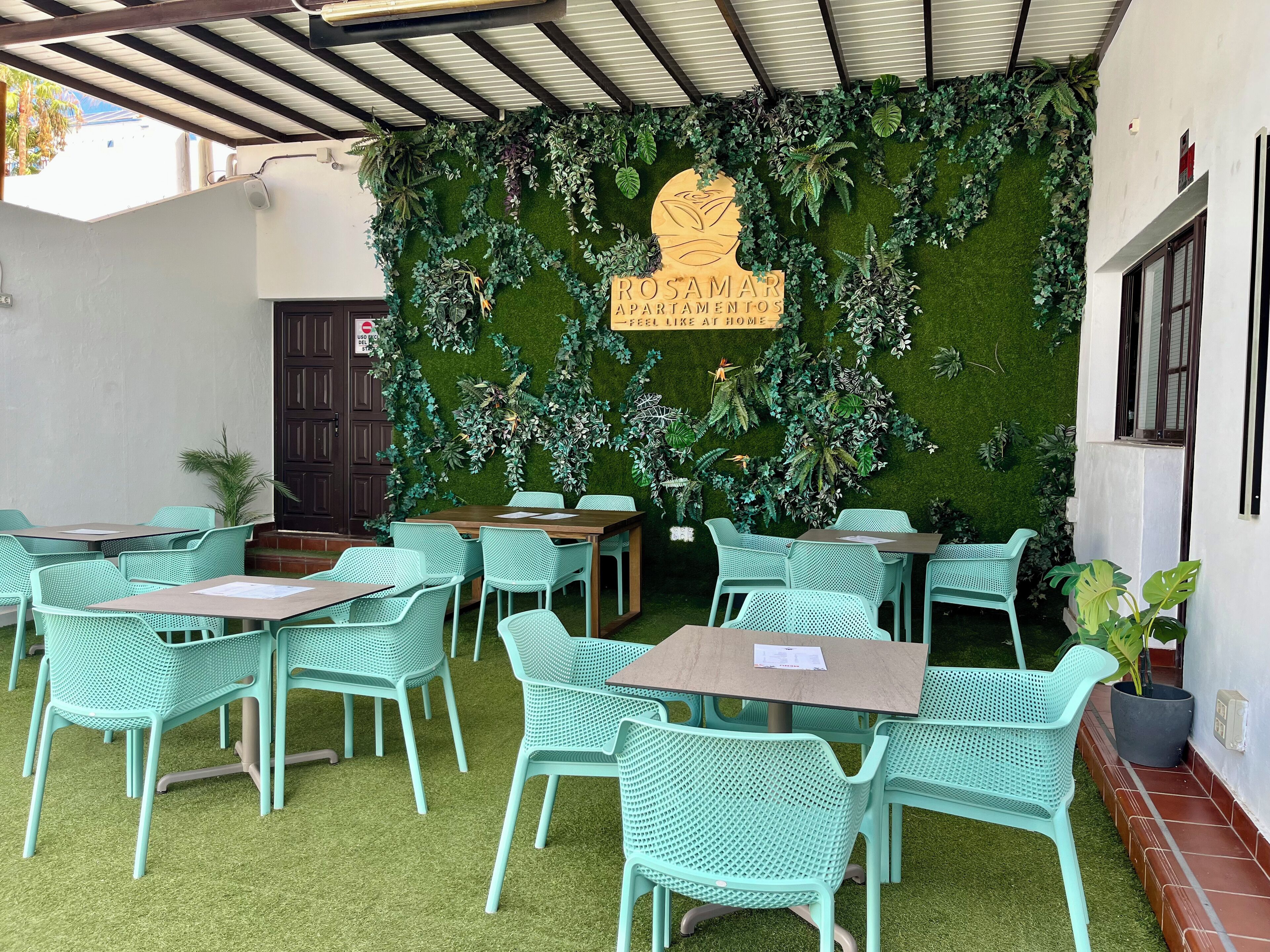A restaurant with outdoor seating, green artificial grass, and a wall decorated with various plants and a sign reading "ROSAMAR APARTAMENTOS - FEEL LIKE AT HOME".
