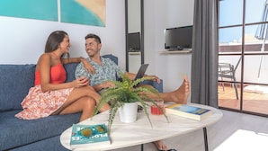 A room with a sofa, a coffee table with a book and a plant, a TV on a stand, and a window with a view of the beach. - Rosamar Resort Lanzarote (Tías)