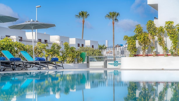 A pool area with lounge chairs and umbrellas, surrounded by palm trees and modern buildings. - Rosamar Resort Lanzarote (Tías)