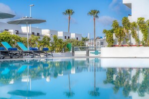 Un área de piscina con sillas de descanso y sombrillas, rodeada de palmeras y edificios modernos.