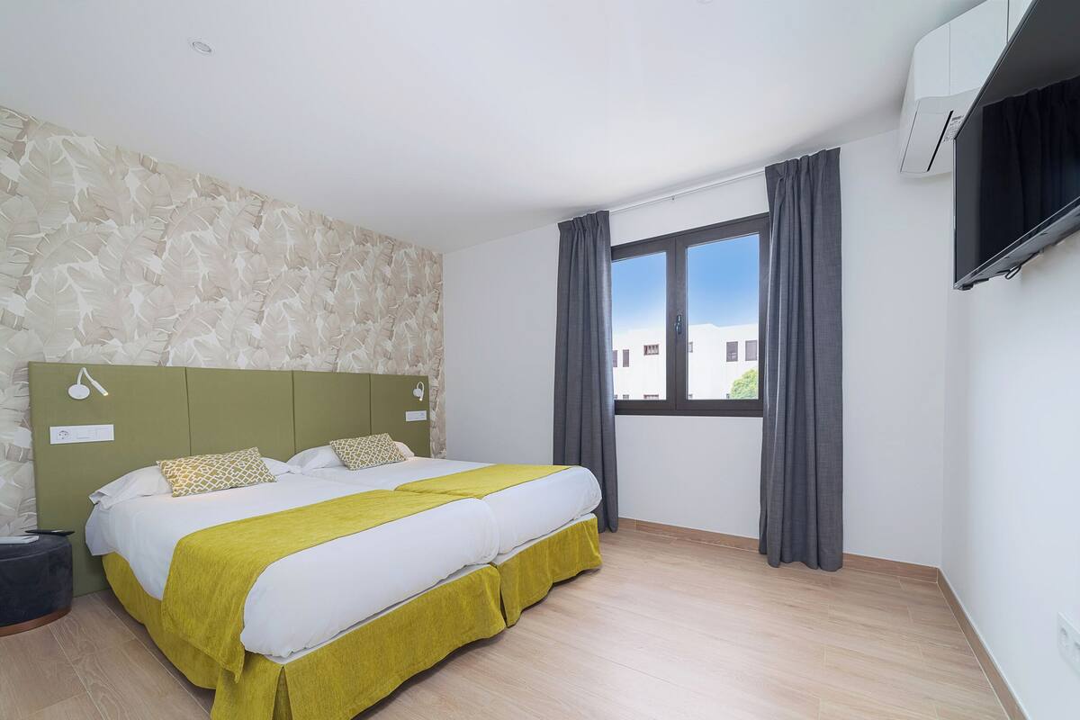 Une chambre d’hôtel moderne avec un grand lit, une tête de lit verte, un téléviseur à écran plat et une fenêtre avec des rideaux.