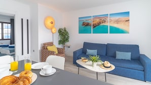 A modern hotel room with a blue sofa, a dining table with breakfast, and a large window with a view of a beach. - Rosamar Resort Lanzarote (Tías)
