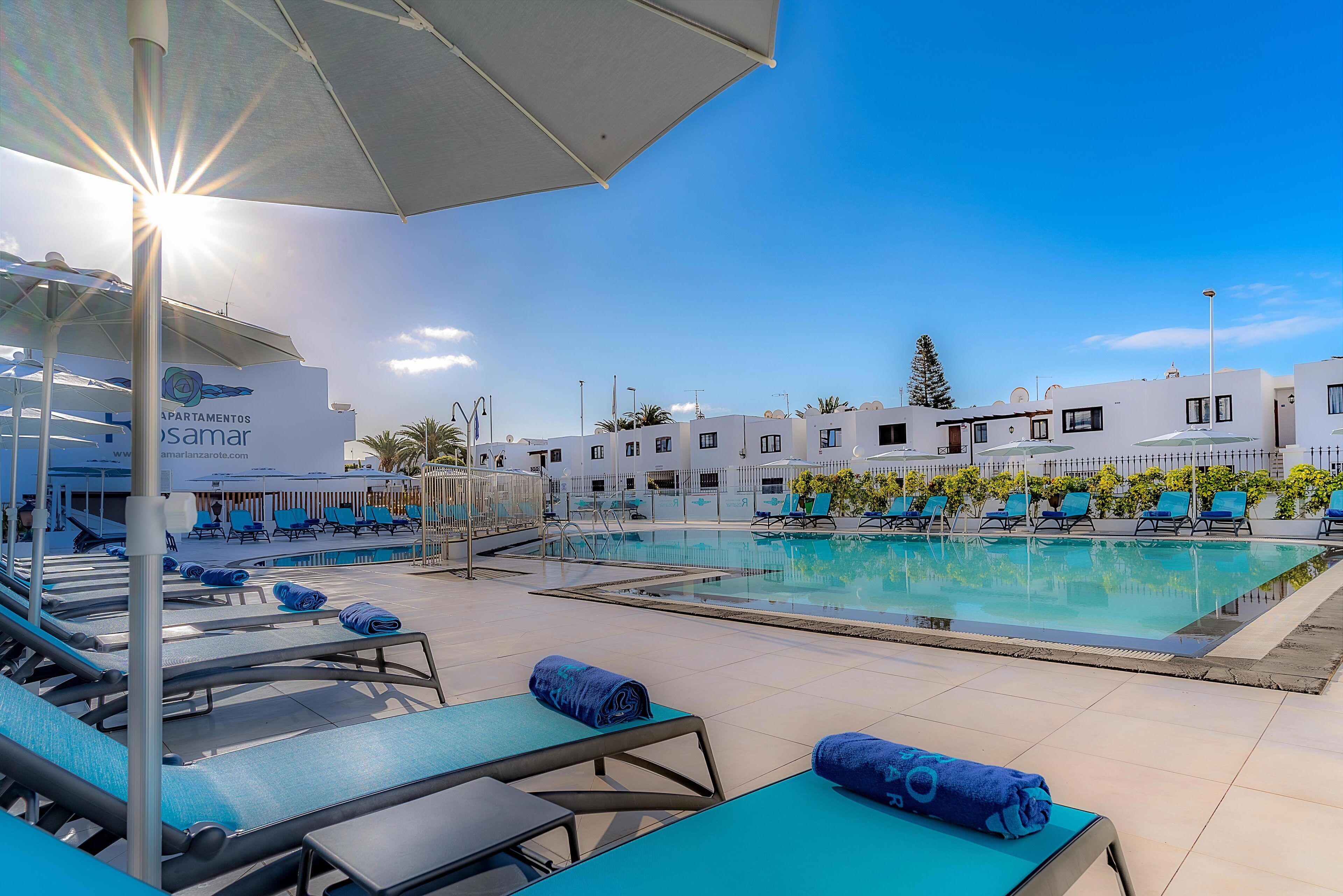 Un área de piscina con tumbonas y sombrillas, un cielo azul despejado y un edificio con un letrero que dice "La Apartamentos Samar".