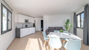 A modern kitchen with white cabinets, a microwave, and a coffee maker. There is a dining area with a white table and light blue chairs. A gray sofa is in the background, and a plant is on the table. - Rosamar Resort Lanzarote (Tías)