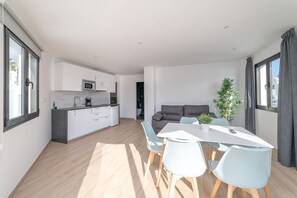 Una cocina moderna con armarios blancos, un microondas y una cafetera. Hay un comedor con una mesa blanca y sillas azul claro. Un sofá gris está al fondo, y hay una planta sobre la mesa.