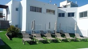 A modern building with a white exterior, a set of sun loungers with umbrellas, and a small garden area. - Apartamentos Miami Playa (San Agustän)
