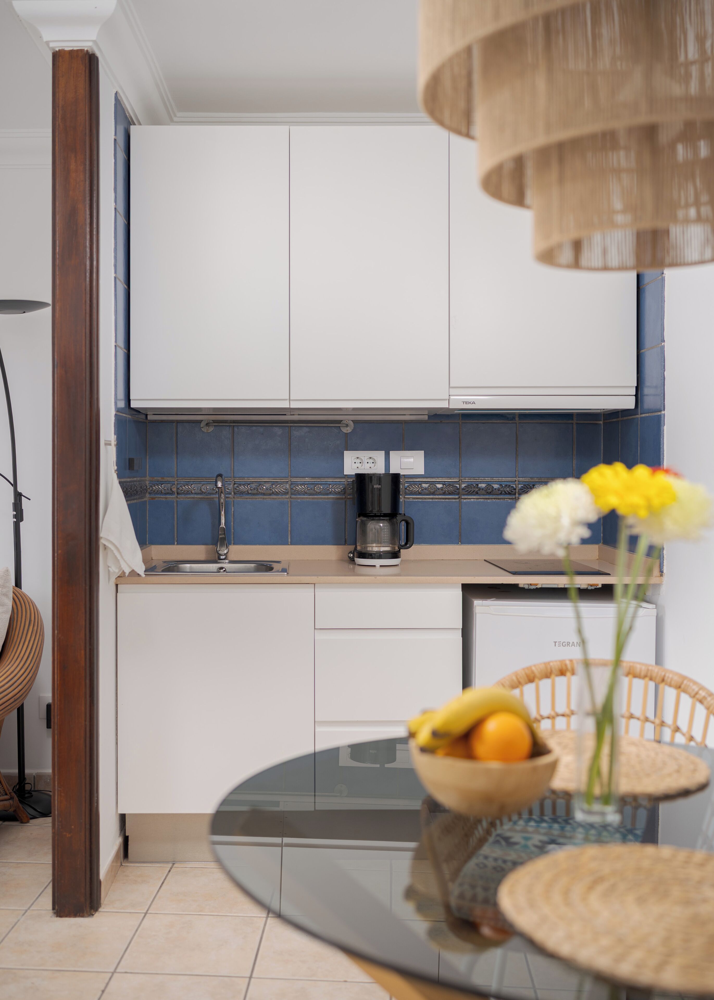 Una cocina moderna con armarios blancos, una cafetera y un comedor con mesa de vidrio y sillas de mimbre.