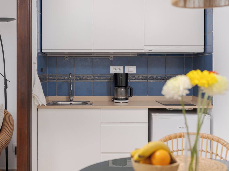 Una cocina moderna con armarios blancos, una cafetera y un comedor con mesa de vidrio y sillas de mimbre.