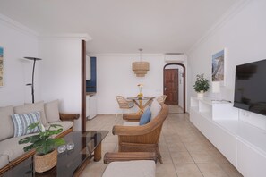 Uma sala de estar com sofá, mesa de centro, cadeiras de vime, uma mesa de jantar com cadeiras, TV e um vaso de plantas.
