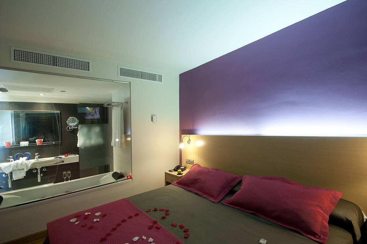 A hotel room with a large mirror, a bed with red pillows, a bedside table with a phone, and a window with a view of the bathroom.