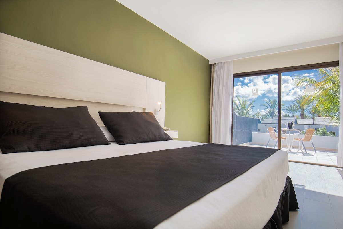 Een hotelkamer met een groot bed, een houten hoofdbord, een balkon met een tafel en stoelen, en uitzicht op palmbomen en de lucht.
