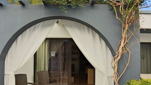 Une pièce avec une entrée dissimulée par un rideau blanc, une table en bois, deux chaises et une vue sur l’intérieur à travers une porte vitrée.