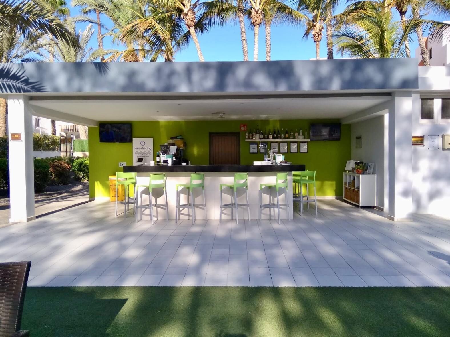 Zona de bar moderna con sillas verdes y una barra, rodeada por una pared blanca y palmeras.