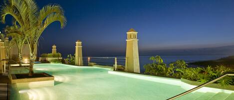 A swimming pool with a view of the ocean at night, surrounded by palm trees and a building with a tower.