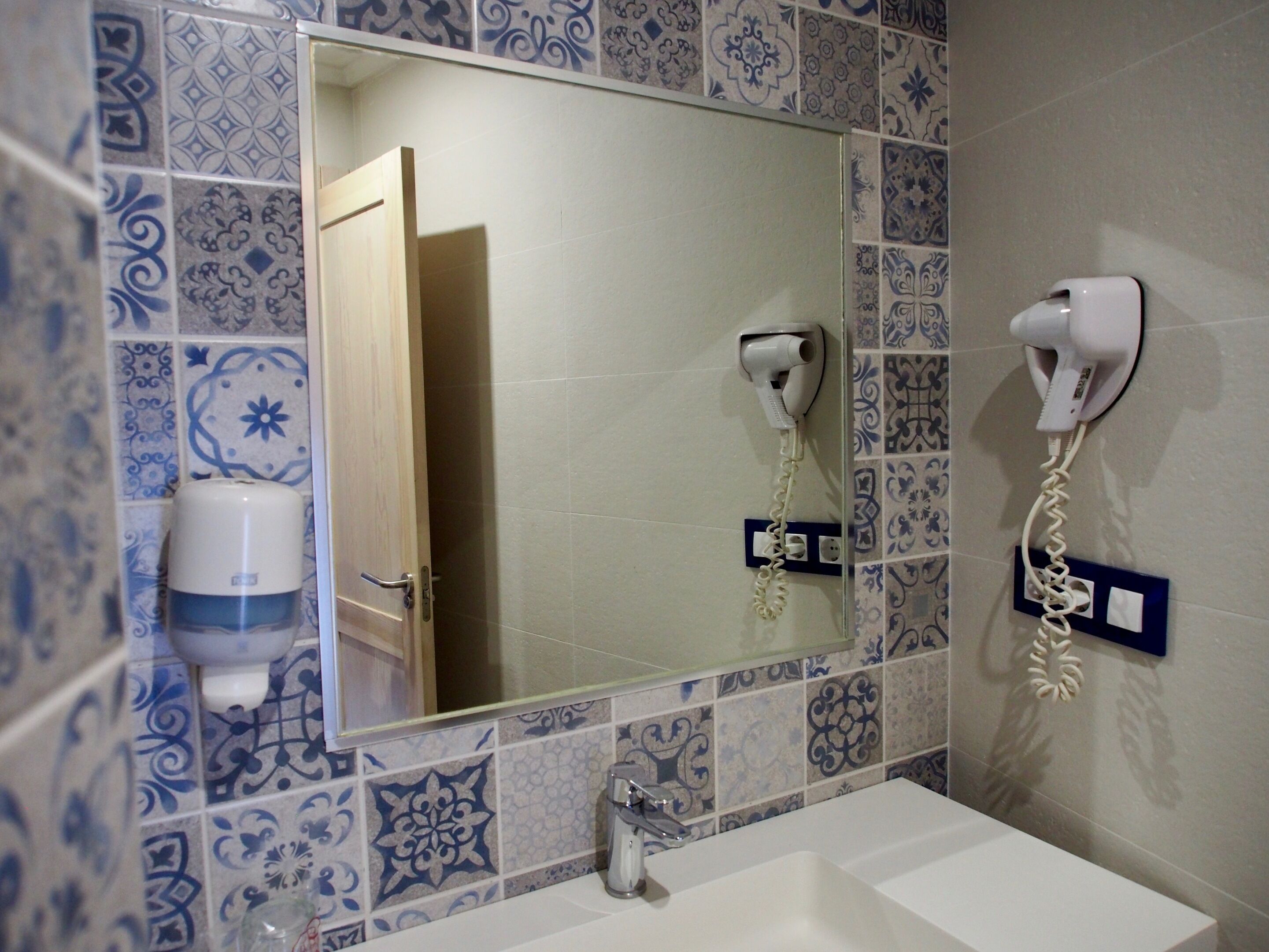 Casa de banho com lavatório, espelho e dois secadores de cabelo fixados na parede.