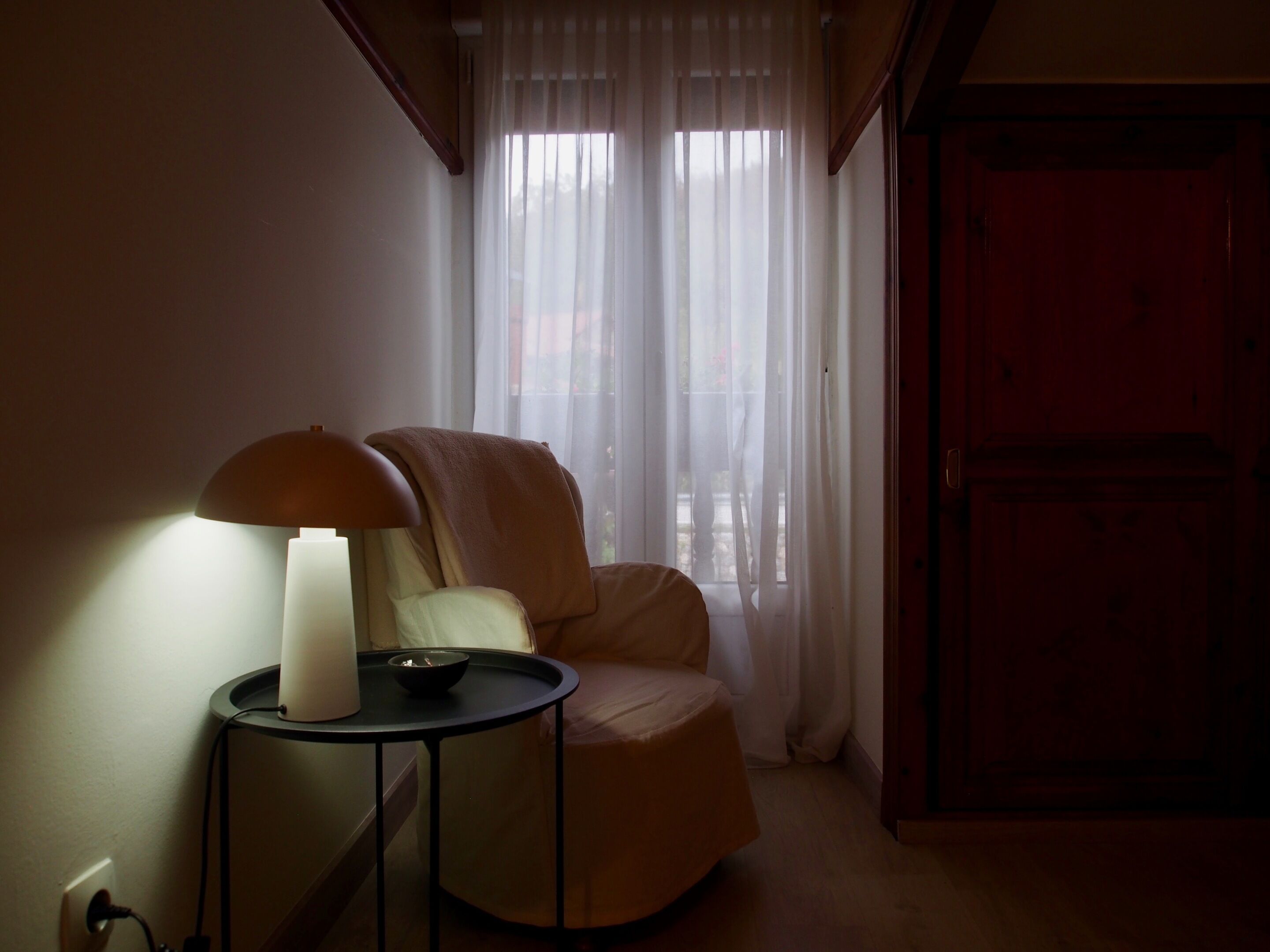 Una habitación con un sillón individual, una mesita con una lámpara y un ventanal con cortinas de gasa.