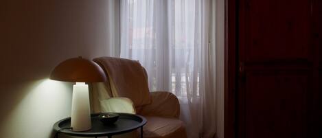 A room with a single armchair, a small table with a lamp, and a window with sheer curtains.