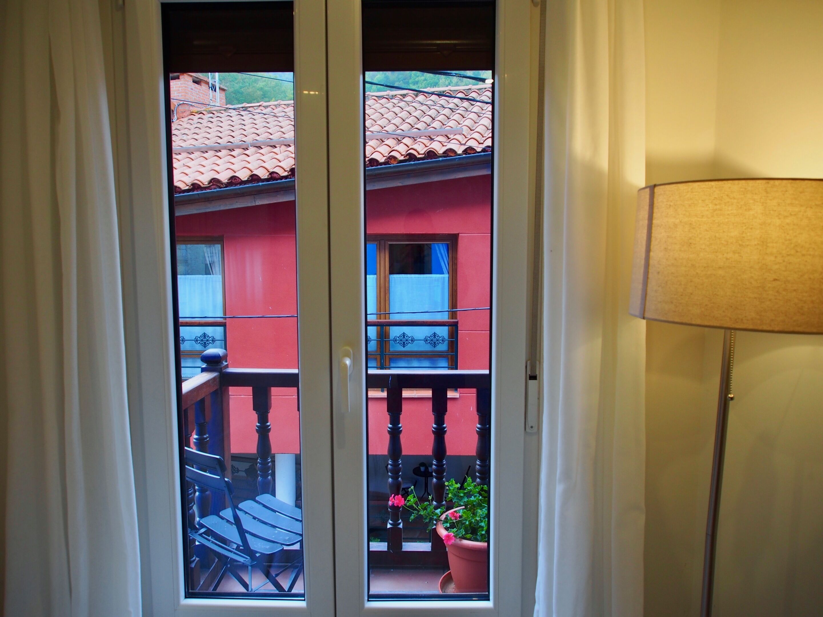 A room with a view of a red building with a tiled roof, a balcony with a chair, and a potted plant.