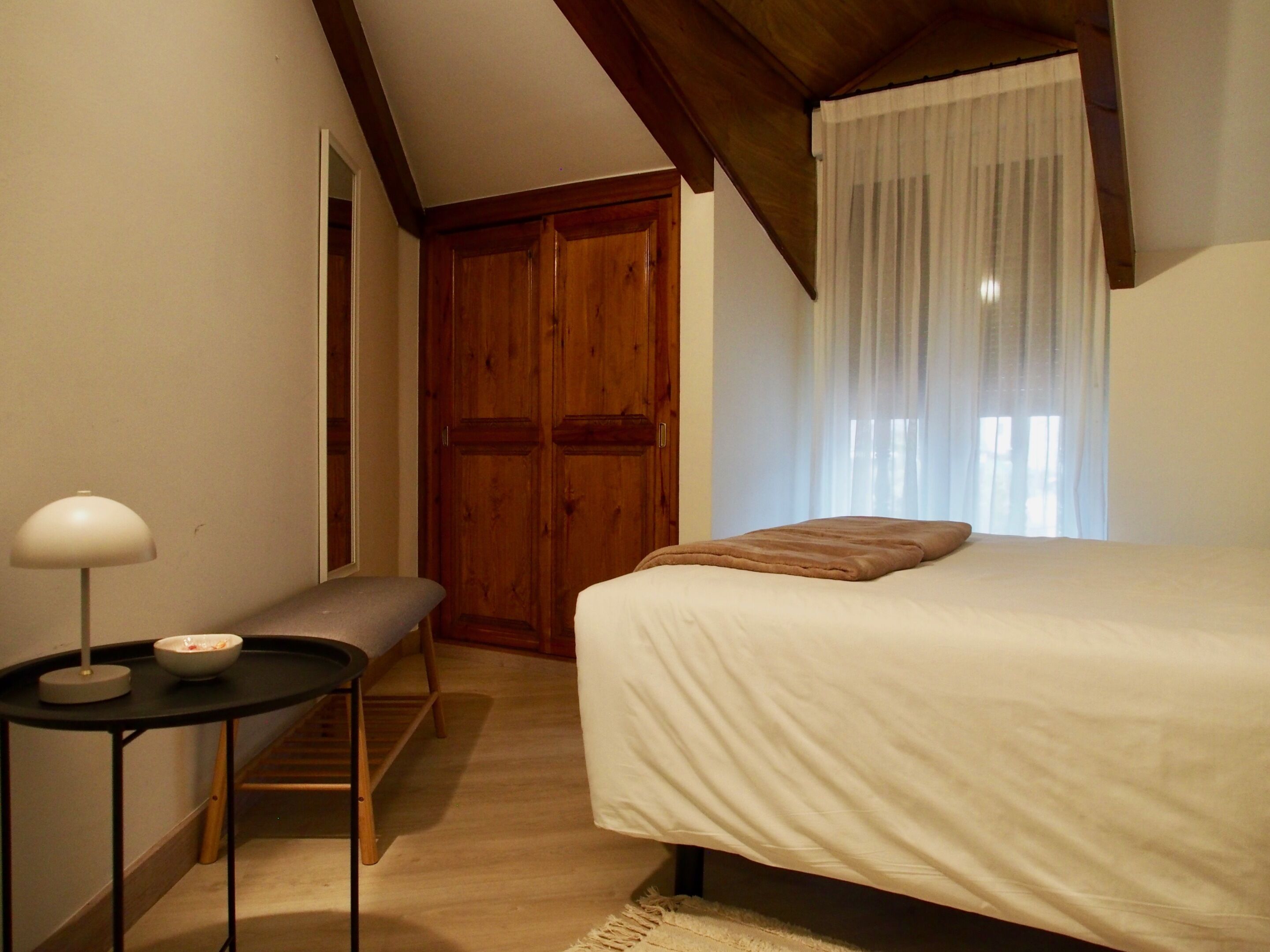 Un dormitorio con una cama, una mesita de noche con lámpara, un banco y un armario de madera.