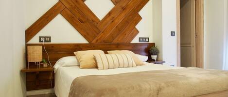 Un dormitorio con cabecera de madera, una cama con ropa de cama color beige, una mesita de noche con lámpara y un florero.
