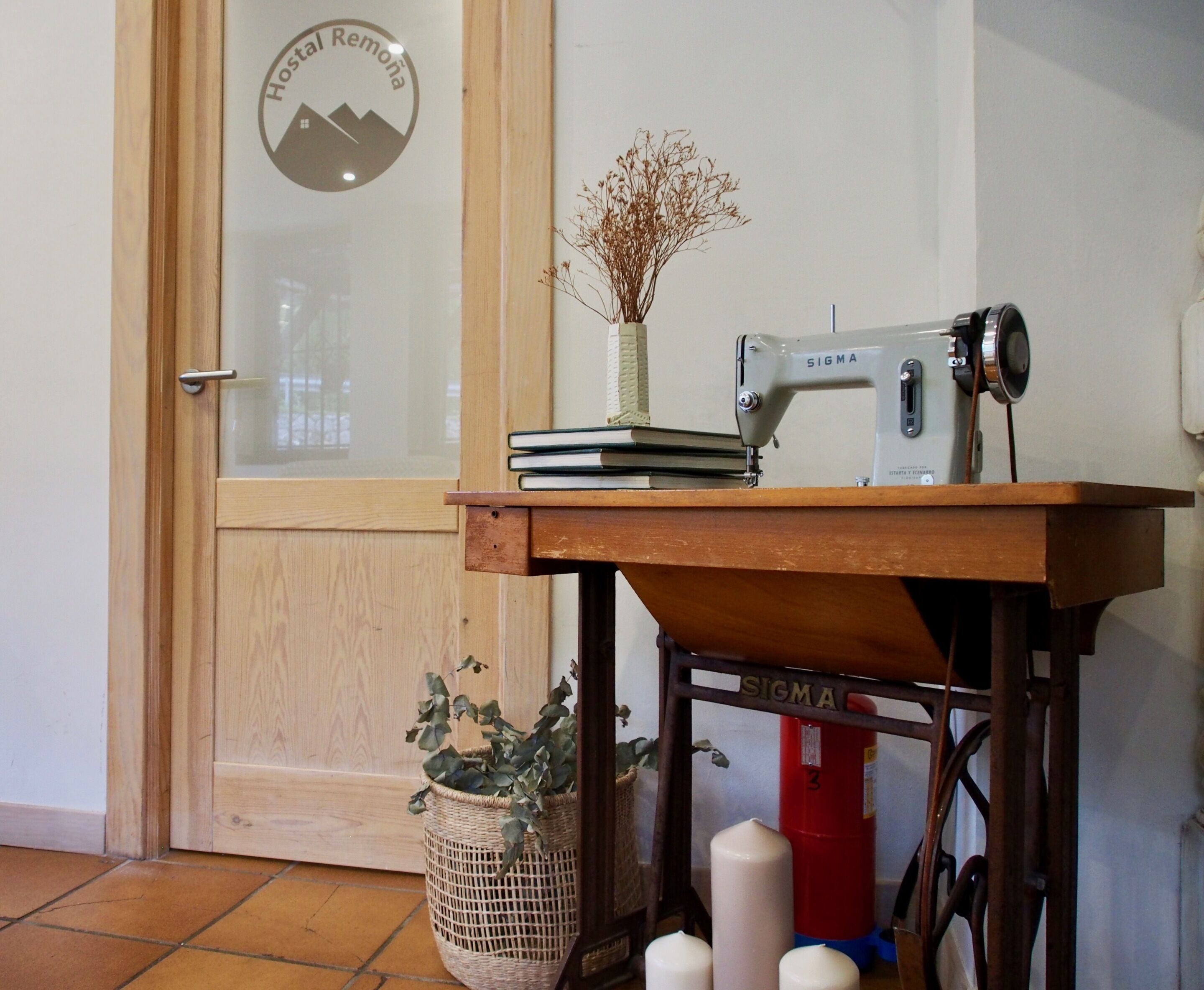 Una máquina de coser sobre una mesa de madera con un florero de plantas secas, una pila de libros y un extintor de incendios al fondo.