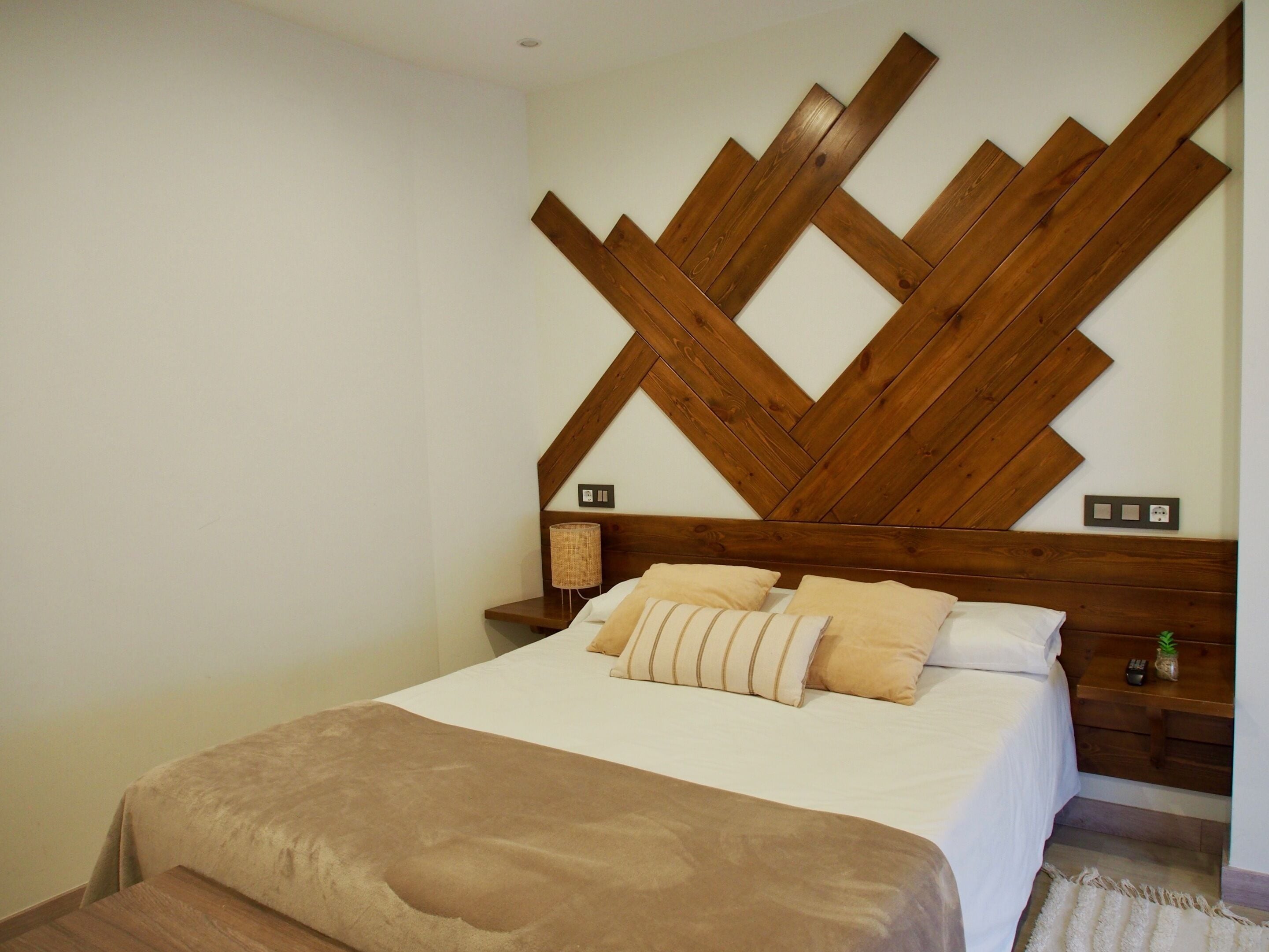 Un dormitorio con cabecera de madera, una cama con manta marrón, dos mesitas de noche con lámparas y una planta pequeña sobre una de ellas.