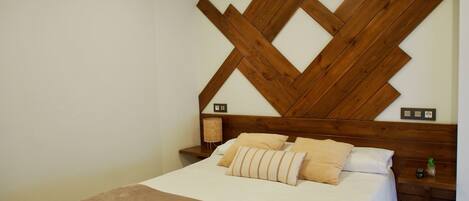 Un dormitorio con cabecera de madera, una cama con manta marrón, dos mesitas de noche con lámparas y una planta pequeña sobre una de ellas.