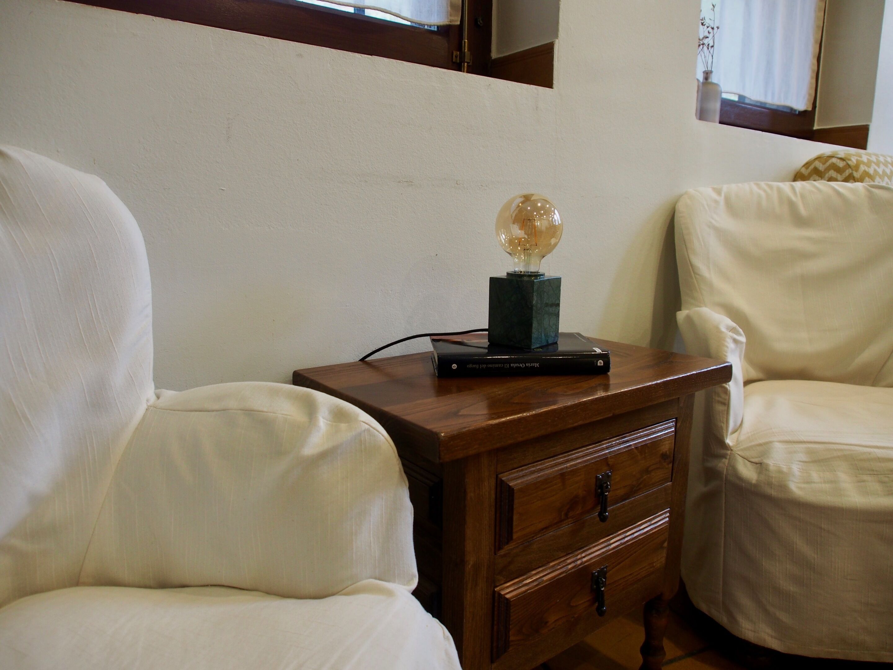 Una mesita de noche de madera con una lámpara y un libro, al lado de un sillón blanco en una habitación con una pared lisa y una ventana.