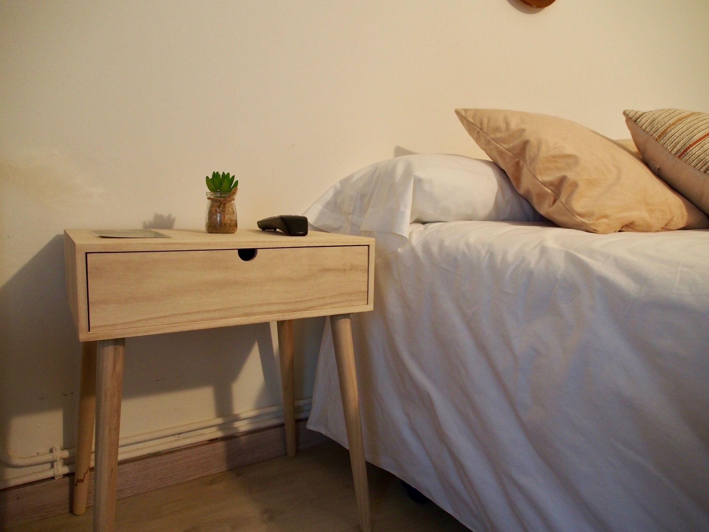 A bedroom with a wooden bedside table, a small potted plant, and a bed with white bedding and beige pillows.