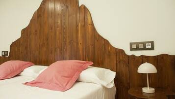 Una cama con cabecera de madera, ropa de cama blanca y almohadas rosas. Hay una mesita de noche con una lámpara y un enchufe.