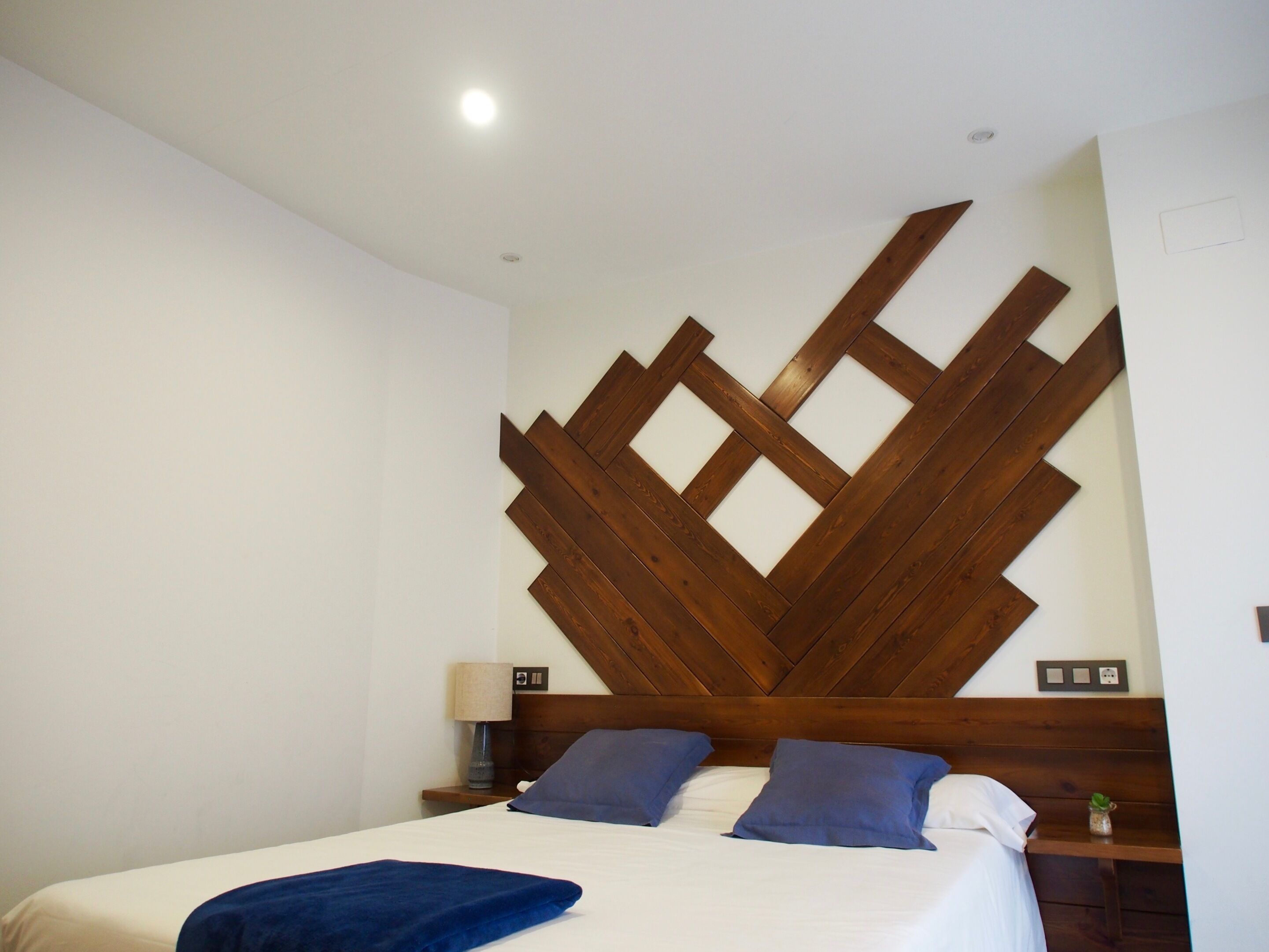 Habitación de hotel con una cama, dos almohadas, una mesita de noche, una lámpara y un cabecero de madera.