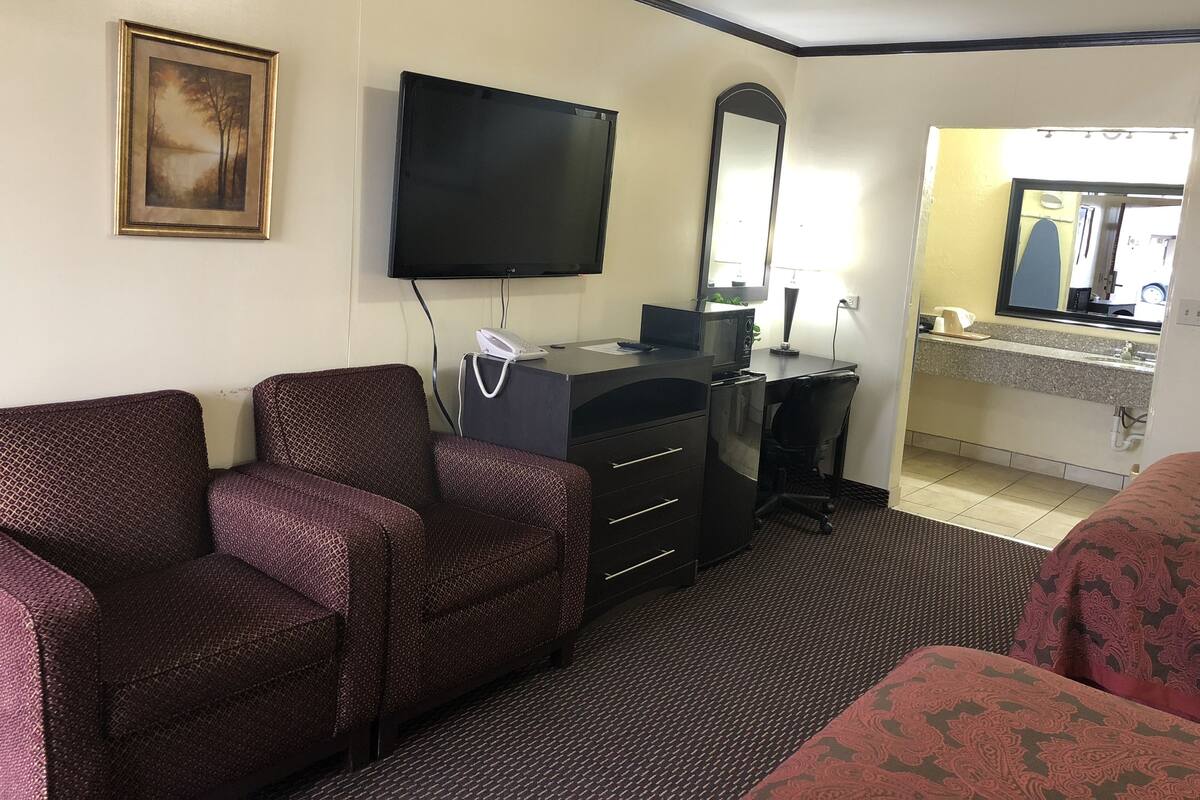 Une chambre d’hôtel équipée d’un téléviseur à écran plat, d’un bureau avec un ordinateur, d’un téléphone et d’un miroir. On y trouve deux fauteuils à motifs et un lit recouvert d’une couvre-lit à motifs.