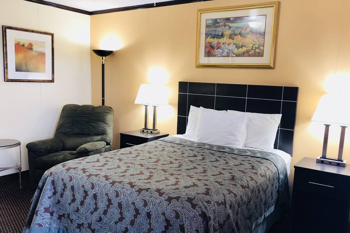 Une chambre d’hôtel avec un lit, une chaise, deux tables de chevet avec des lampes et deux tableaux encadrés accrochés aux murs.
