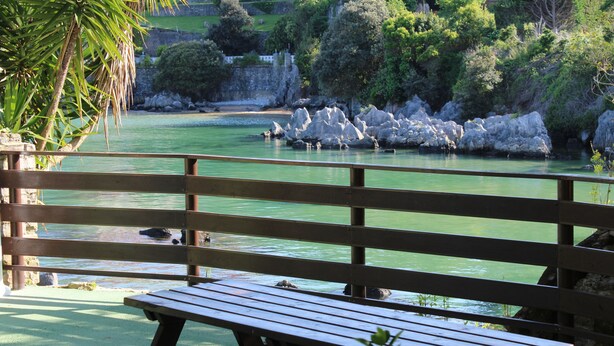 Un banco de madera con vista a un cuerpo de agua tranquilo, con rocas y follaje al fondo.