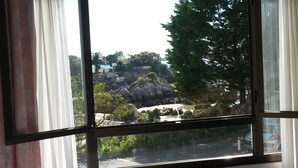 Vista a través de una ventana con cortinas, mostrando un paisaje rocoso con árboles y una carretera.