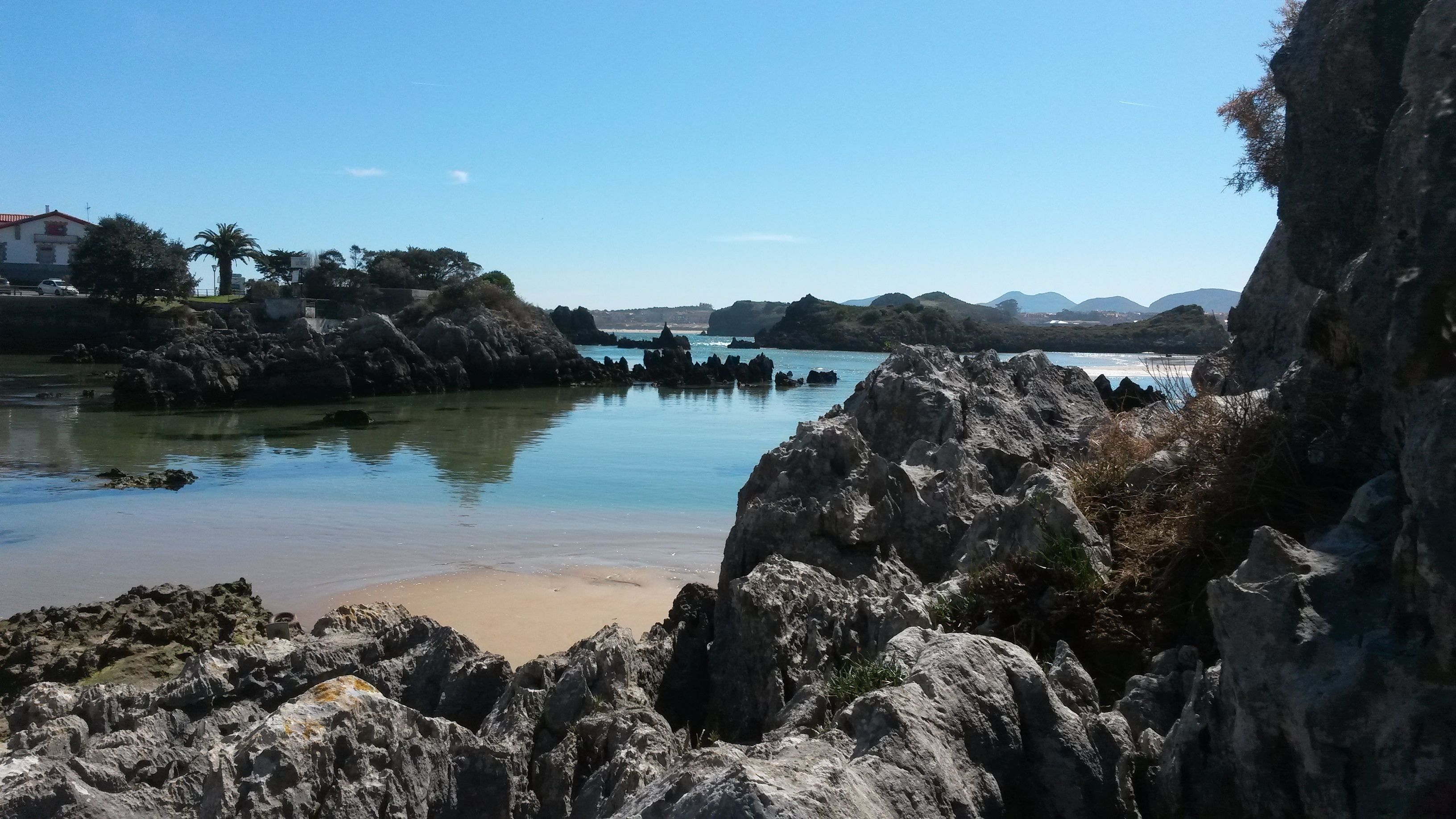 A coastal landscape with rocky formations, calm water, and distant mountains.