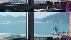 A room with a large window overlooking a beach and mountains. There are two potted plants and a tray with glasses on the windowsill. - Hotel Risco (Laredo)