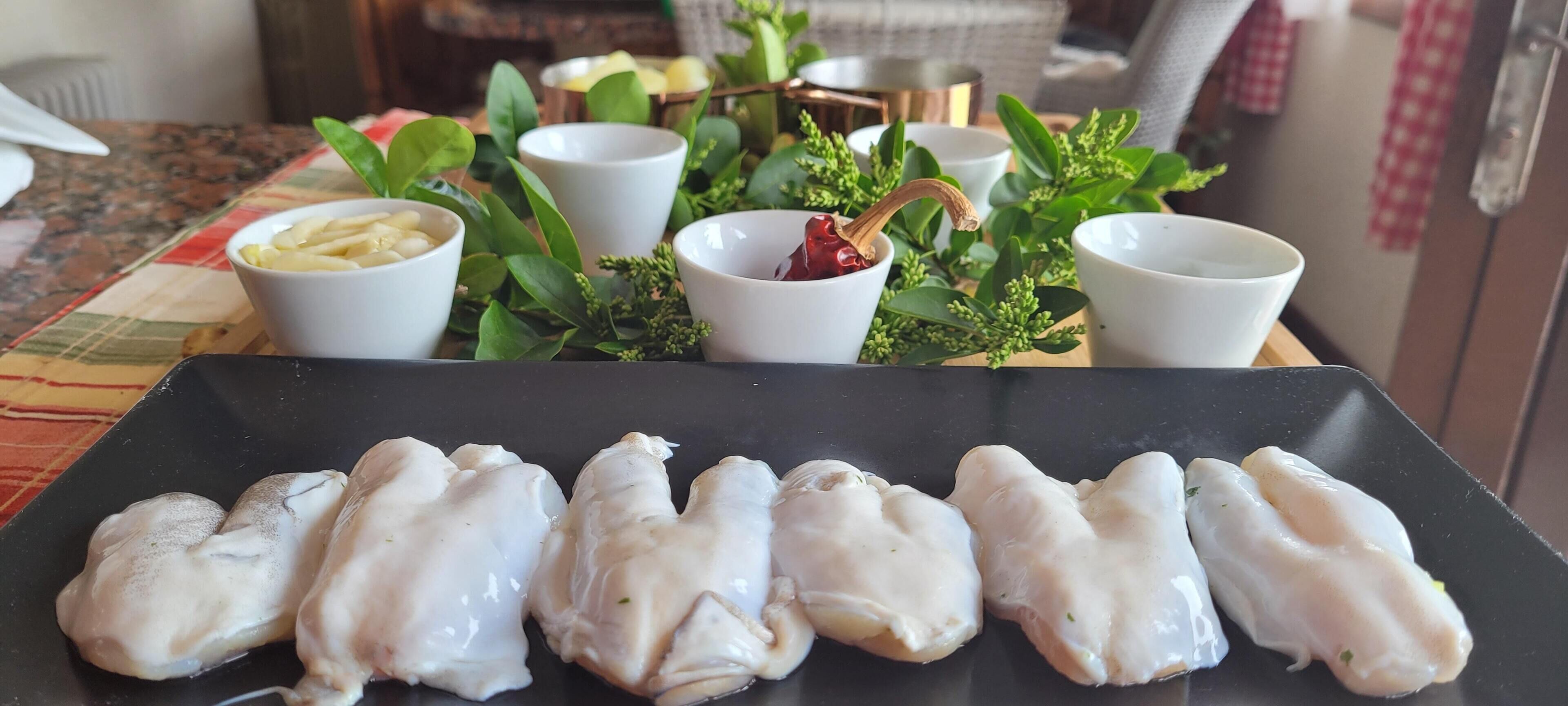 Une table avec une assiette noire contenant du poulet cru, des bols blancs remplis de divers ingrédients et des plantes vertes à feuilles.