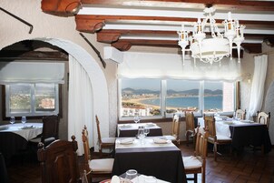 A dining area with tables set for two, wooden chairs, and a view of the sea through large windows.