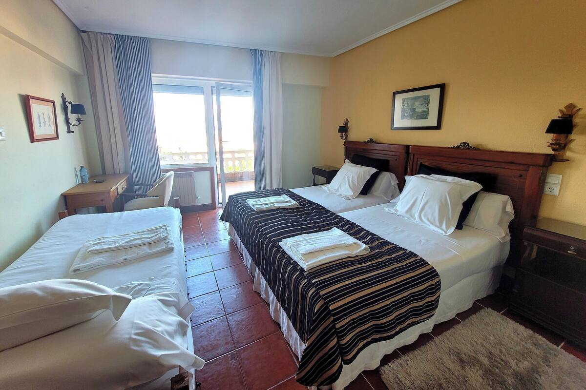 Ein Hotelzimmer mit zwei Betten, einem Schreibtisch, einem Stuhl, einem Fenster mit Vorhängen und Blick nach außen.