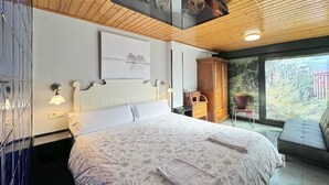 Une chambre dotée d’un grand lit, d’un plafond en bois, d’une vue sur la verdure grâce à une porte vitrée, et d’une œuvre d’art accrochée au mur au-dessus du lit.