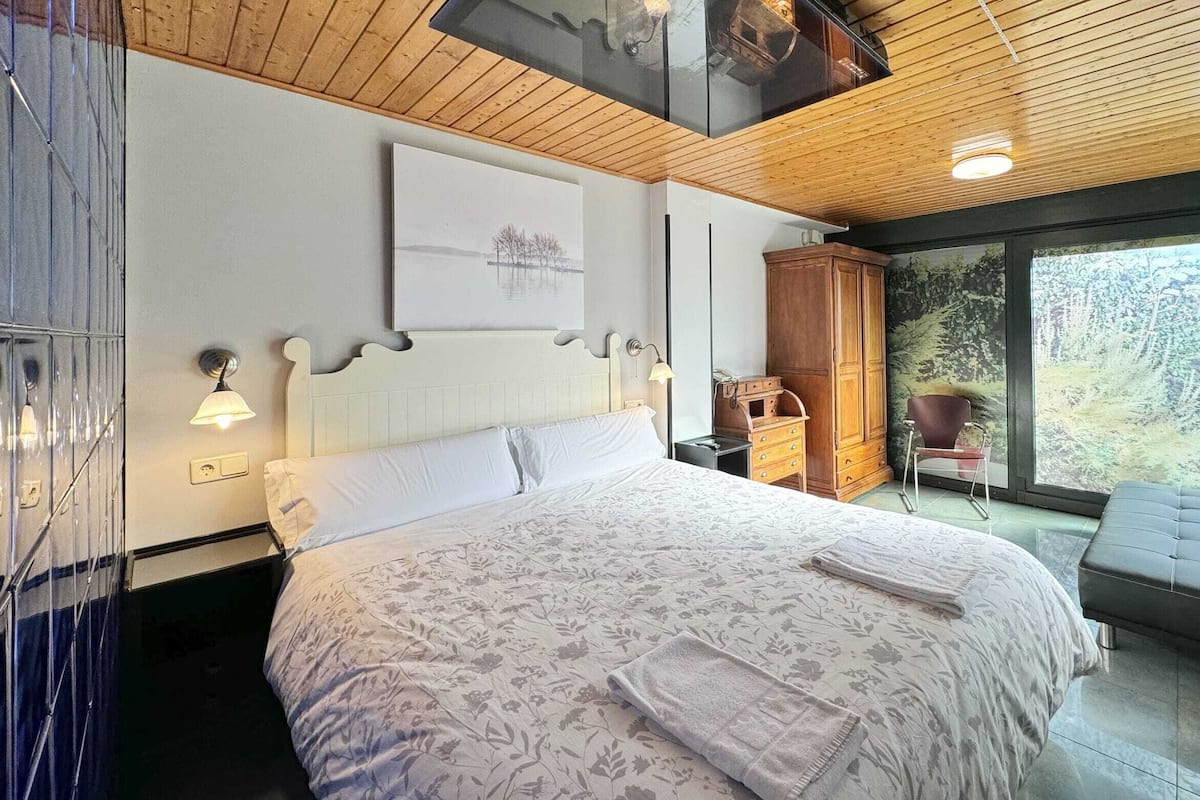 Ein Schlafzimmer mit einem großen Bett, Holzboden, Blick ins Grüne durch eine Glastür und einem Kunstwerk über dem Bett.