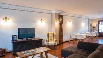 A spacious living room with a television, a large armchair, a sofa, and a dining area with chairs and tables.