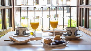 Una mesa de desayuno con dos vasos de jugo de naranja, un plato de pasteles y vista a un balcón con barandilla.
