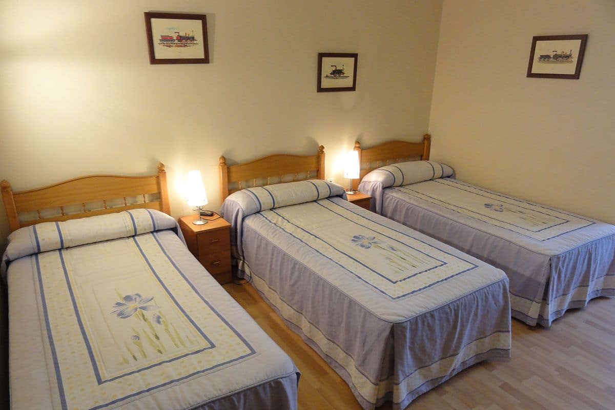 A hotel room with three single beds, each with a wooden headboard and a bedside table with a lamp.